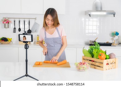Asian beautiful woman, long hair, wearing a pink t-shirt and striped apron standing, slicing carrots on the table at modern white kitchen, and recording her cooking video using a mobile phone. - Shutterstock ID 1548758114