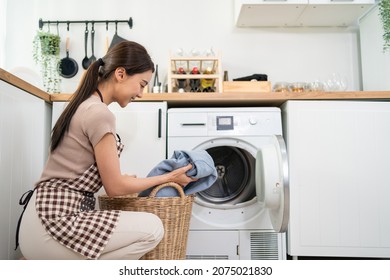 Asian Beautiful Woman Put Dirty Clothes To Washing Machine In House. Attractive Girl Wear Apron Sit On Floor, Feel Happy To Loads Laundry In Washer Appliance At Home. Domestic-House Keeping Concept.