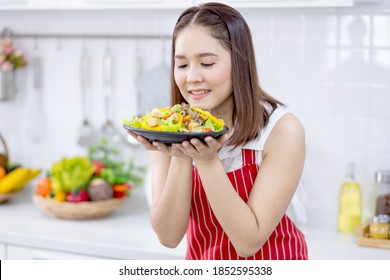 Asian Beautiful Woman With Red Apron Hold Plate With Filled In Food And Action Of Good Smell In Kitchen And Look To Camera With Smiling. Concept Of Happiness Of Cooking In Their House.