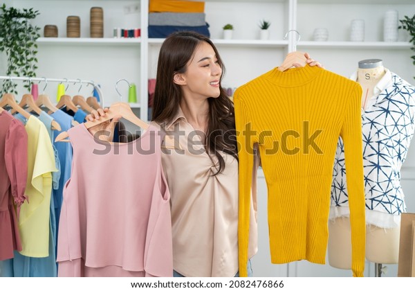 Asian beautiful woman sell cloth product online\
live streaming at home. Young female use mobile phone video call\
shows goods to customer and present detail. Remote buying and\
purchase shopping concept