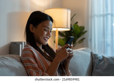 Asian Beautiful Woman Sit On Sofa And Chat On Mobile Phone At Night. Attractive Young Happy Girl Spend Leisure Time At Home, Feel Relax And Enjoy Communicate And Discuss On Smartphone In Living Room.