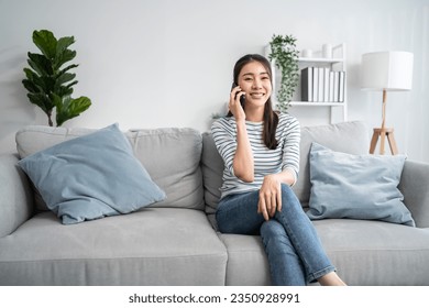 Asian beautiful woman sit on sofa and talk on mobile phone in house. Happy attractive young girl spend leisure time at home, feel relax and enjoy communicate and discuss on smartphone in living room. - Powered by Shutterstock