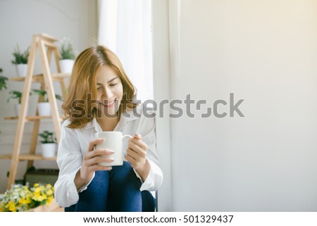 Asian beautiful woman sitting by the window with cup of tea or coffee in her hands on the light background,flare light