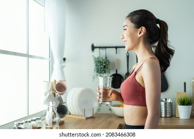 Asian beautiful woman in sportswear drink water after exercise at home. Young thirsty active sport girl takes a sips of clean mineral natural in cup after workout for health care in kitchen in house.