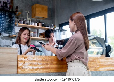 Asian Beautiful Woman Use Mobile Phone To Pay With Contactless Payment And Service By Barista Girl Or Coffee Maker In Coffee Shop. Concept Of New Normal Payment And Technology Support Small Business.