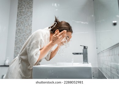 Asian beautiful woman washing her clean face with facial foam and water. Attractive female in bathrobe washing face for healthy beauty treatments and skin care then looking at the mirror in bathroom.