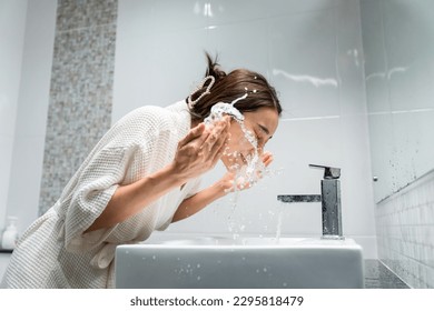 Asian beautiful woman washing her clean face with facial foam and water. Attractive female in bathrobe washing face for healthy beauty treatments and skin care then looking at the mirror in bathroom. - Powered by Shutterstock