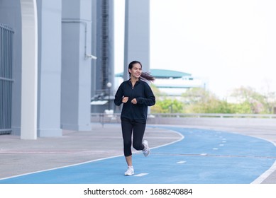 Asian Beautiful Young Woman Black Suit With Happy Running Or Jogging On Blue Running Track In The Day Time. Exercise, Training, Fitness, Workout, Sport, Lifestyle And Healthy Concept. 