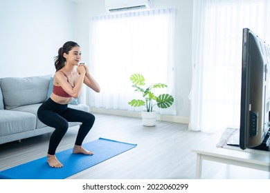 Asian Beautiful Young Woman Stay Home, Doing Aerobic Exercise At Home. Attractive Girl Doing Lockdown Activity, Workout By Squatting, Follow Instructions Video From Online Trainer For Health In House.