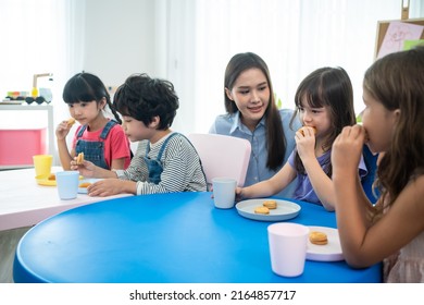 Asian Beautiful Young Woman Teacher Serving Snack To Kid At Schoolroom. Attractive Instructor Master Female Give The Student A Break And Serve Cookie And Drink To Kindergarten Pre School In Classroom.