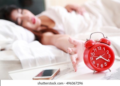 Asian Beautiful Young Woman Waking Up Late. Sleepy Woman Long Black Hair With Nightdress Is Reaching For Red Alarm Clock Is Ringing Next To Mobile Phone At Table Near White Bed In Bedroom On Morning.