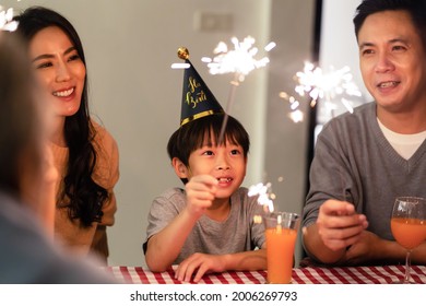 Asian Big Happy Family, Parent Play Sparkler With Young Kid Together. Young Little Boy Child Sit Between Father And Mother On Dinner Table, Enjoy Birthday Party At Night. Activity Relationship Concept