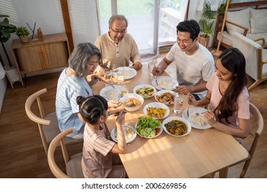 Asian Big Happy Family Spend Time Have Lunch On Dinner Table Together. Little Kid Daughter Enjoy Eating Foods With Father, Mother And Grandparents. Multi-Generation Relationship And Activity In House