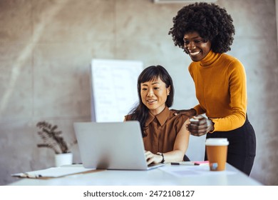 An Asian and a Black businesswoman engage in a collaborative effort, reviewing content on a laptop in a well-lit workspace, dressed in smart casual attire. - Powered by Shutterstock