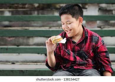 Asian Boy Eating Muffin Sandwich On A Green Concrete Bench