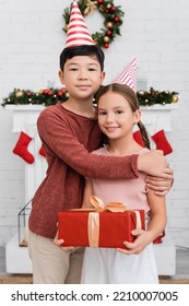 Asian Boy Hugging Friend In Party Cap Holding Gift Box Near Blurred Christmas Decor At Home