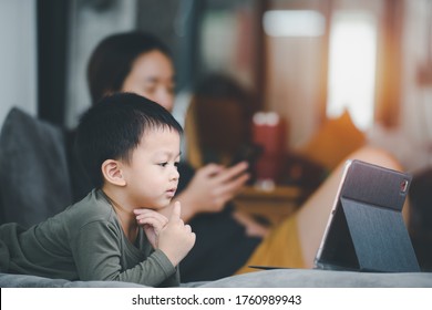 Asian Boy Looking At The Tablet Screen Attentively. Overstimulated Children Concept. Too Much Screen Time. 3 Years Boy Watching Videos While Tv Is Working. Internet Addiction Concept.