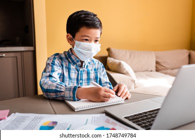 Asian Boy In Medical Mask Writing And Studying Online With Laptop At Home During Quarantine