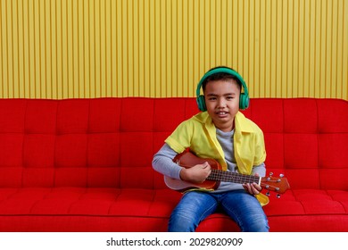 Asian Boy On Grey Sweater, Yellow Shirt, Blue Jeans, Wearing Green Earphone And Sit On Red Sofa Of Striped Stage To Concentrate On Singing And Performing Rock Music Show By Playing Small Guitar