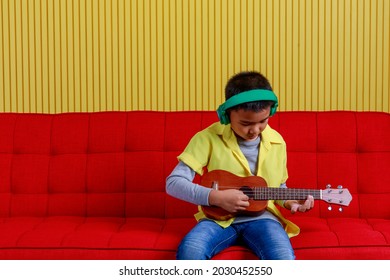 Asian Boy On Grey Sweater, Yellow Shirt, Blue Jeans, Wearing Green Earphone And Sit On Red Sofa Of Striped Stage To Concentrate On Singing And Performing Rock Music Show By Playing Small Guitar