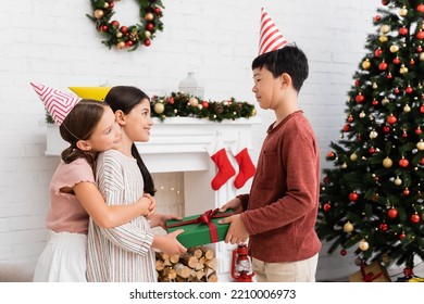 Asian Boy In Party Cap Giving Present To Cheerful Friend Near Christmas Decor On Fireplace At Home