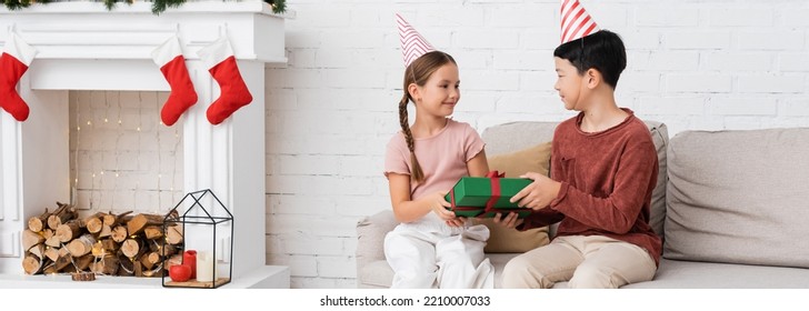 Asian Boy In Party Cap Giving Present To Friend During Birthday Party Near Christmas Decor At Home, Banner