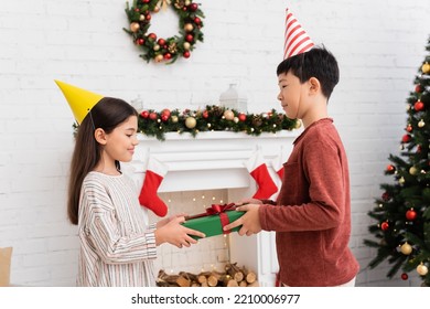 Asian Boy In Party Cap Holding Gift Box Near Smiling Friend And Christmas Decor At Home