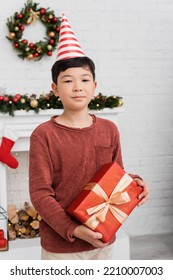 Asian Boy In Party Cap Holding Present And Looking At Camera Near Blurred Christmas Decor At Home