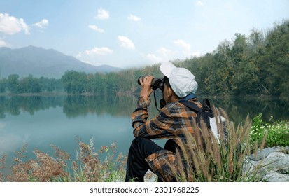 Asian boy in plaid shirt wears white cap, holding binoculars, sitting on reservoir ridge during summer vacation and birdwatching activity, soft and selective focus, nature study and hobby concept.