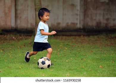 Asian Boy Play Football In The Park. Kid Kick Ball In Grass Field.