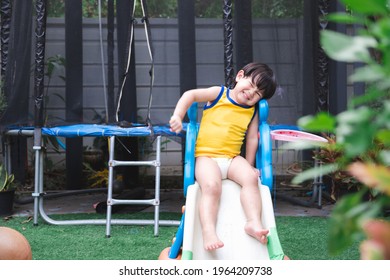 Asian Boy Plays Small Slide In Front Yard House. Child Smile Sweet. Children Is Wearing Yellow Sports Shirt. Baby Wear Disposable Diapers. Kid Is In Good Mood. Happy Toddler Is 2-3 Years Old.