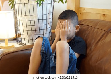 Asian boy sits on the sofa with his hands covering his face. Concepts of shy, insensitive, sad, afraid, depressed, ashamed, crying,worried.                               