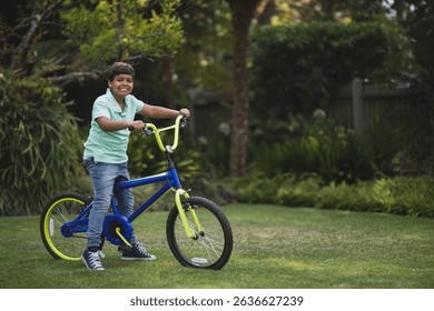 Asian boy smiling while straddling blue bicycle with neon yellow forks on garden lawn. Playful, outdoor, vibrant, youth, leisure, nature, lifestyle