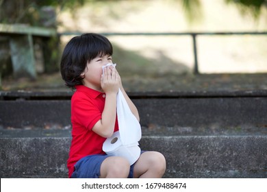 Asian Boy Snezzing While Holding Tissue Paper Roll.Out Of Stock Concept On Corona Virus Pandemic.Little Boy Playing With Tissues In Outdoor.Red Shirt Little Preschooler Bored.