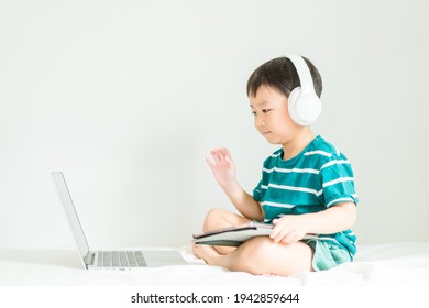 Asian Boy Student Wear Wireless Headphone Study Online With Video Call Teacher, Toddler Boy Child Online Learning Listen English Class.write Notes Look At Laptop Home.Covid19 Coronavirus.Education.