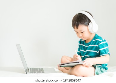 Asian Boy Student Wear Wireless Headphone Study Online With Video Call Teacher, Toddler Boy Child Online Learning Listen English Class.write Notes Look At Laptop Home.Covid19 Coronavirus.Education.