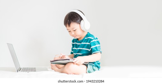 Asian Boy Student Wear Wireless Headphone Study Online With Video Call Teacher, Toddler Boy Child Online Learning Listen English Class.write Notes Look At Laptop Home.Covid19 Coronavirus.Education.