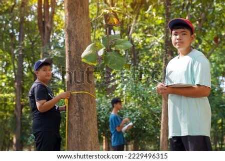 Asian boy taking notes on trees measured by friends to calculate the carbon storage of trees. Concept Learning outside the classroom about the environment, global warming, carbon credits.             