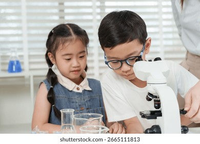 Asian boy using microscope during elementary science class while girl watches beside in classroom with lab glassware and assistance from female teacher during hands-on learning activity