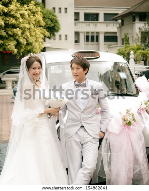 Asian Bride in white\
dress and asian groom in white suit posing at in front of their\
vintage wedding car.