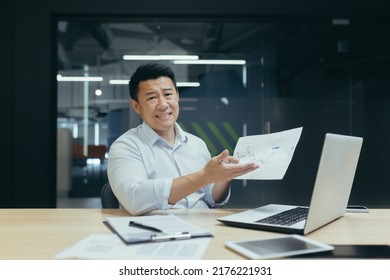 An Asian Broker Is Disappointed With The Company's Financial Indicators, Looks At The Camera Not Happy, Shows A Document With A Negative Income Schedule, The Man Works In The Office.