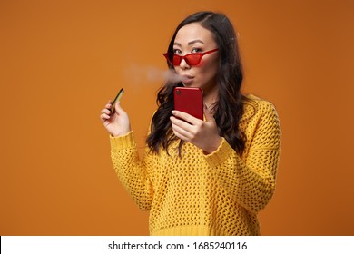Asian brunette girl in glasses and with phone in her hand smoking vape