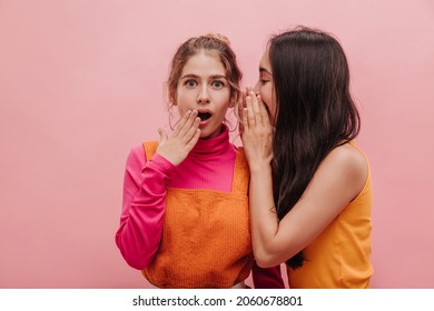 Asian Brunette Woman Whispers Secret To Her Friend From Europe, Surprising Her On Pink Background With Place For Text. Image Of Two Women From 20s Discussing Gossip With Each Other.