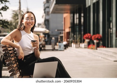 Asian brunette woman with wide smile sitting on bench looks at camera with coffee in her hands on city street. Girl manager in non-office is engaged in additional work at laptop.