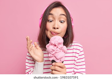Asian brunette young woman looks at appetizing ice cream licks lips eats yummy frozen dessert listens music via headphones dressed in striped jumper isolated over pink background. Mmm how tasty - Shutterstock ID 1983107591