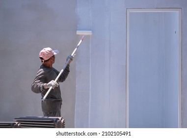 Asian builder worker using long handle roller brush to painting primer white color on concrete wall inside of house construction site