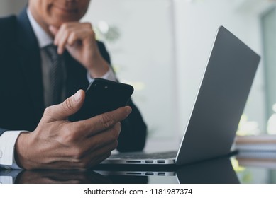 Asian Business Man In Black Suit Using Smart Mobile Phone And Working On Laptop Computer, Checking Internet Mobile Apps In Modern Office, Close Up. Online Working,  Business And Technology Concept