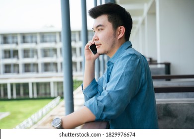 Asian Business Man In Blue Collar Shirt Talking On The Phone At Hotel Balcony.