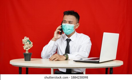 Asian business man wearing mask called with laptop on table isolated red background - Powered by Shutterstock