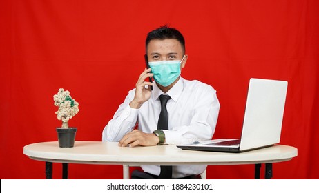 Asian business man wearing mask called with laptop on table isolated red background - Powered by Shutterstock